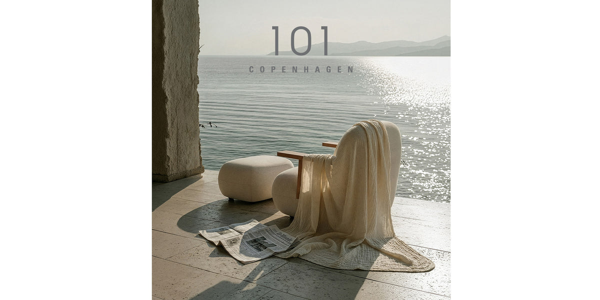 A styled interior scene with a cream draped Toe chair with wooden armrest, ottoman, and blanket on a sunlit wooden floor beside a large window overlooking water. Large overlaid text reads '101 Copenhagen'.