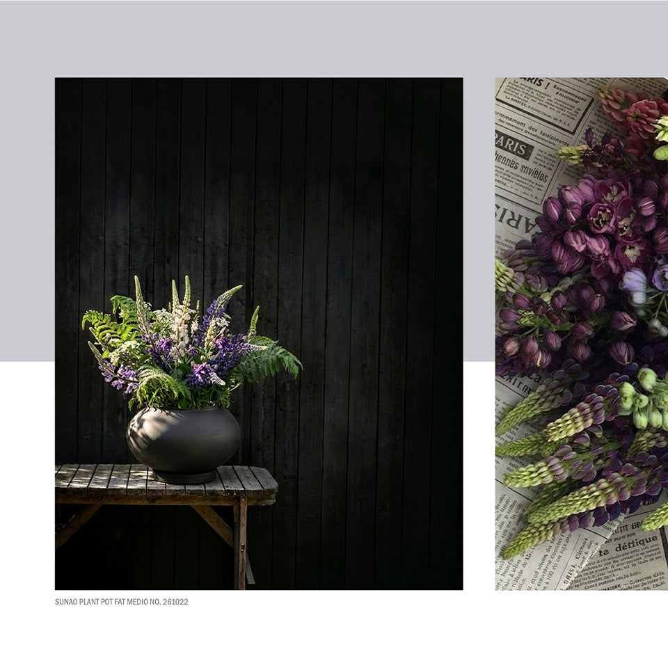 A floral arrangement in a Sunao plant pot displayed on a wooden table against a dark wood plank wall, shown alongside a close-up detail of the bouquet with purple flowers and green foliage on a newspaper-patterned surface.