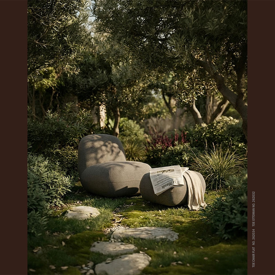 A modern curved Toe lounge chair in a mossy garden setting, with an Ottoman draped in a light beige throw and a newspaper on its seat. The scene is framed by dense green foliage and trees with a dark brown border on the left and right.