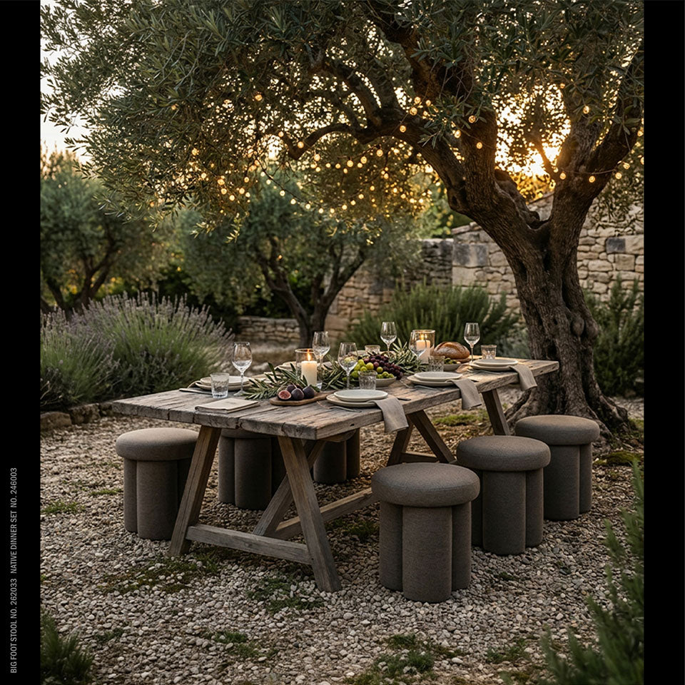 Outdoor dining setup under a large tree with string lights, featuring a rustic wooden table set with Native plates, wine glasses, candles, and bread, surrounded by Big Toe stools in a garden courtyard.