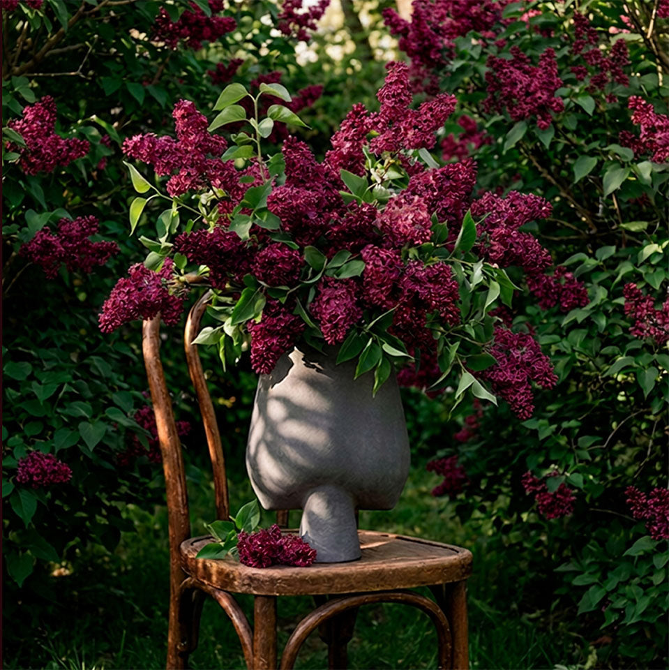 A gray sculptural Sphere vase shaped like a stylized head, filled with dense clusters of deep purple lilac flowers and green leaves, displayed on a rustic wooden chair in a garden setting.