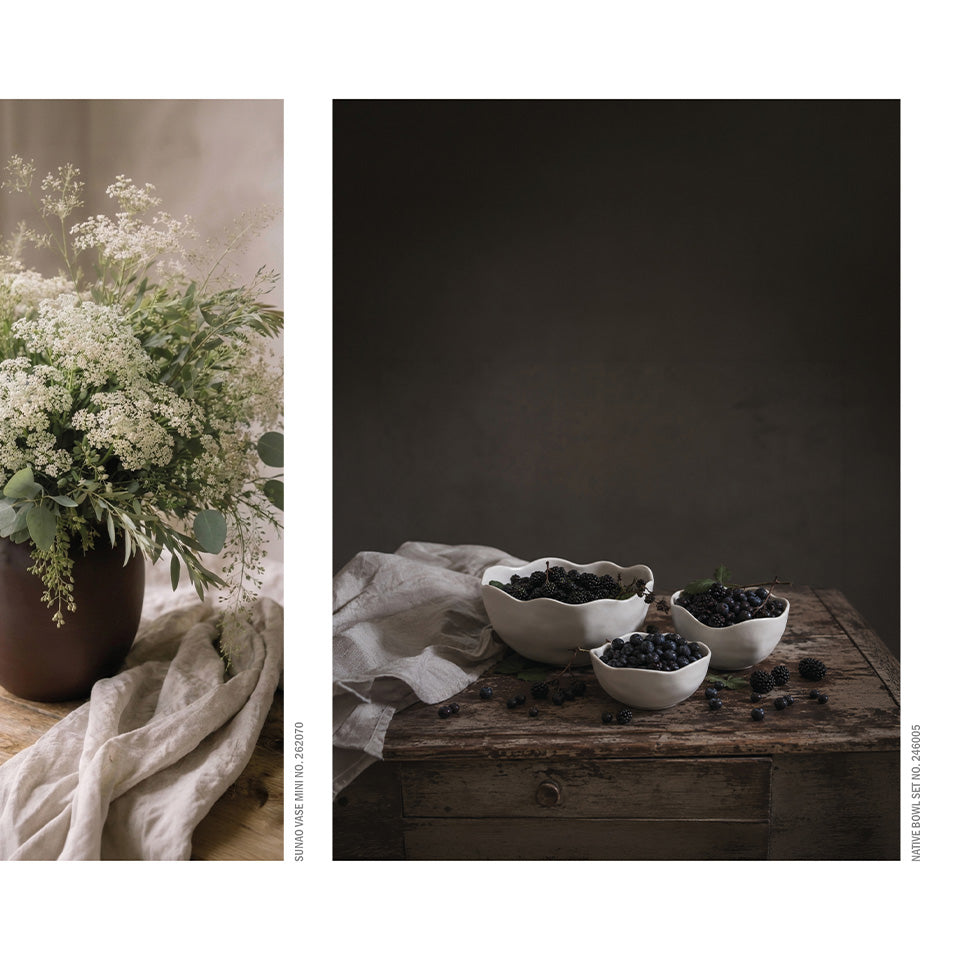 A split photographic composition: the left panel shows a floral arrangement in a dark vase with white blossoms and greenery draped over a light fabric; the right panel shows three white Native bowls filled with dark blueberries on a rustic wooden table against a dark brown background.