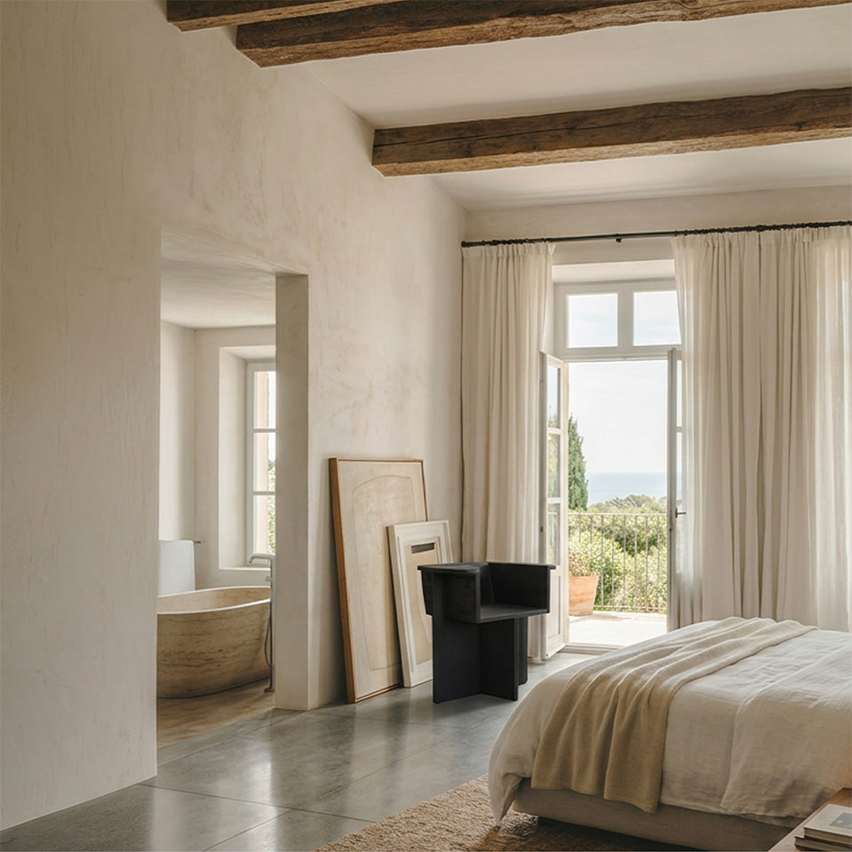 A minimalist bedroom interior with a bed dressed in cream bedding, tall white curtains, exposed wooden ceiling beams, and a large window opening to a garden. A Brutus Dining chair i Coffee black oak. A bathtub is visible in an adjacent room through an open doorway.