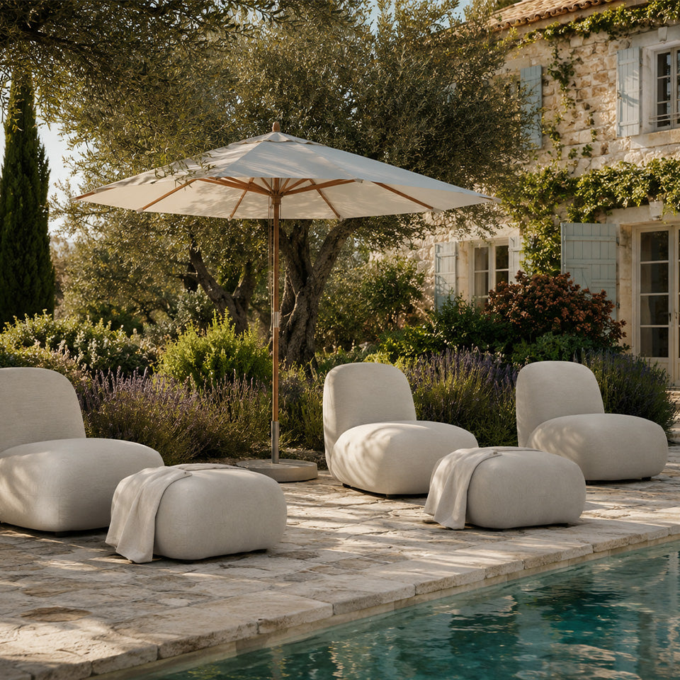 Outdoor patio furniture set by a swimming pool, featuring four Toe Chair and ottoman a large beige patio umbrella, set against a stone building and lush garden.