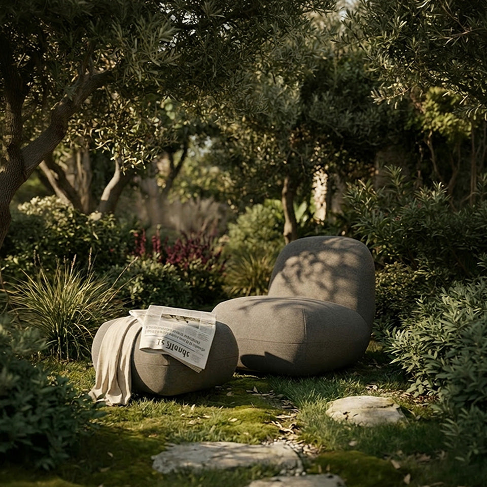 Two modern, organic-shaped outdoor Toe lounge chair and Ottoman in a mossy garden setting, with one draped in a cream throw blanket and a newspaper.