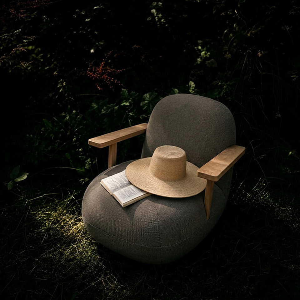 A dark gray upholstered Toe lounge chair with light wood arms sits outdoors on grass in a wooded setting. A beige wide-brim straw hat and an open book rest on the chair.