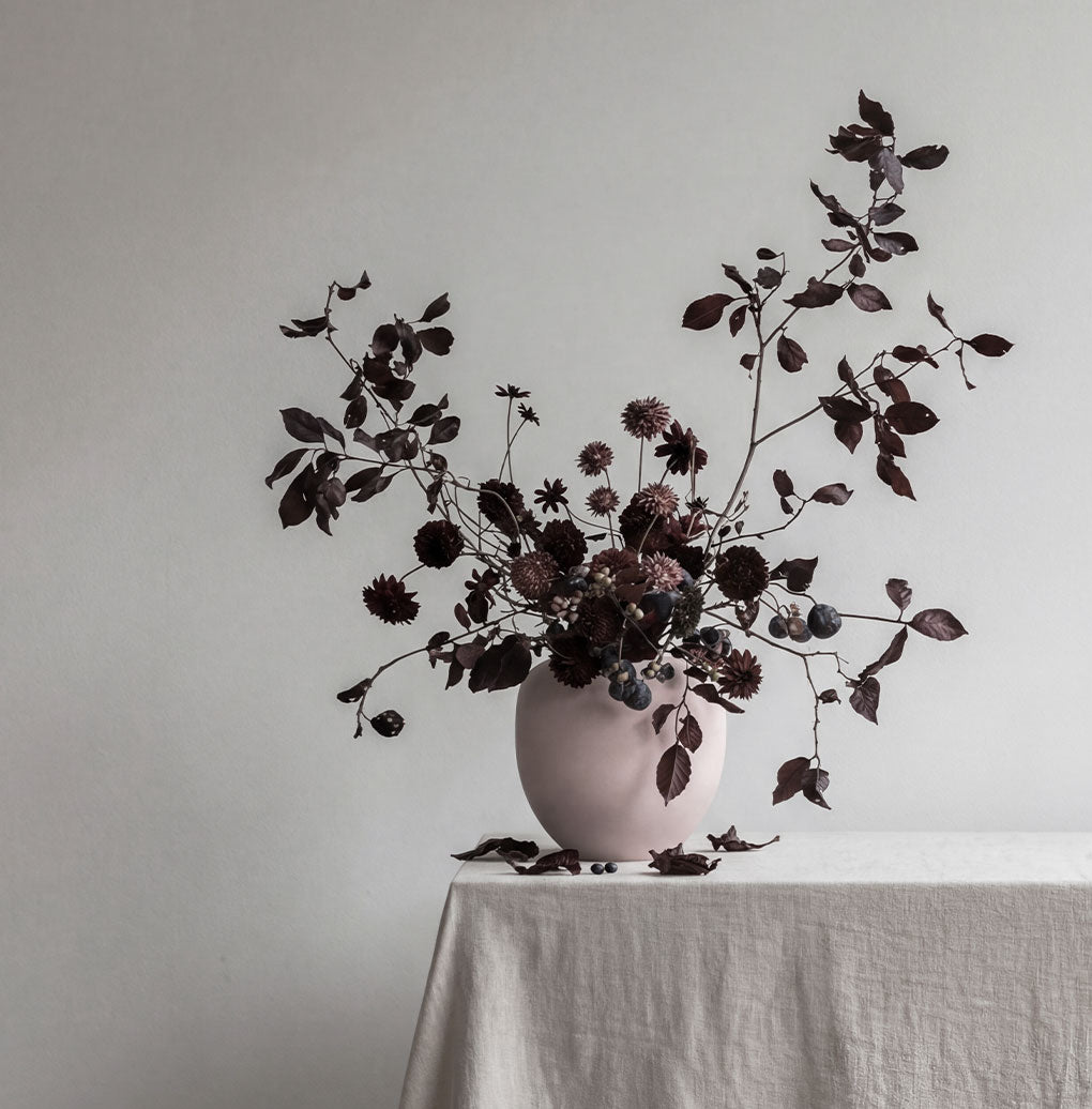 Rose Sunao vase with dark flowers on a tablecloth-covered table against a plain wall