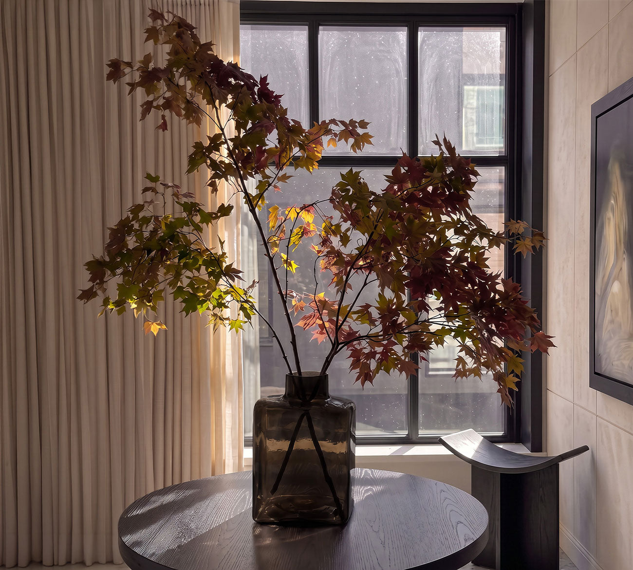 A Shin Vase Big i Smoked Forest glass holding a tall arrangement of red and green maple leaves, placed on a round dark table in a modern interior room with a window and beige curtains.