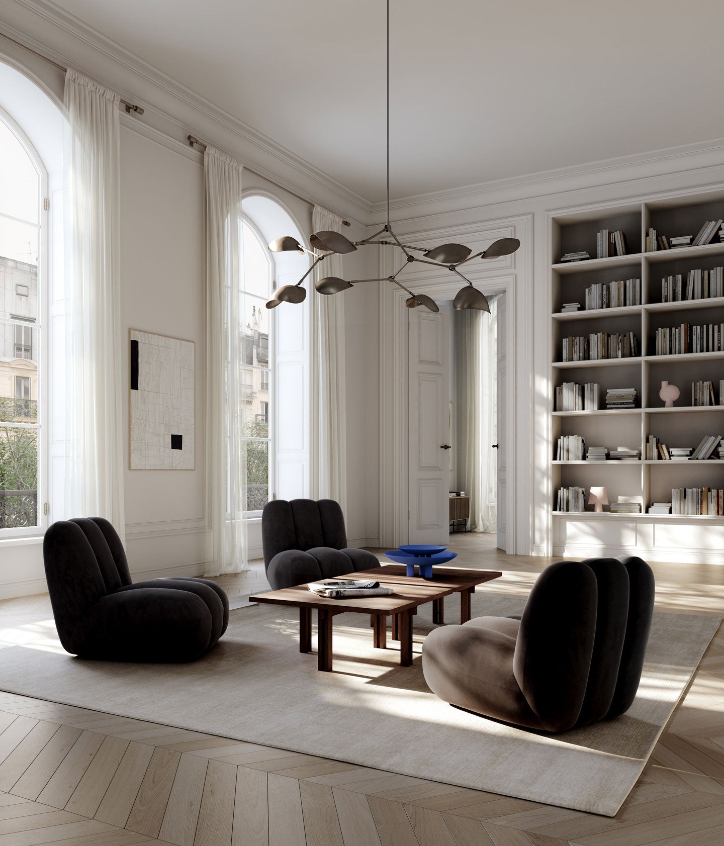 A modern living room with three sculptural dark Toe chairs arranged around a wooden Plinth coffee table on a large cream rug. The room features high arched windows with white drapes, a built-in bookshelf filled with books, and a contemporary multi-arm Stingray chandelier hanging from the ceiling.