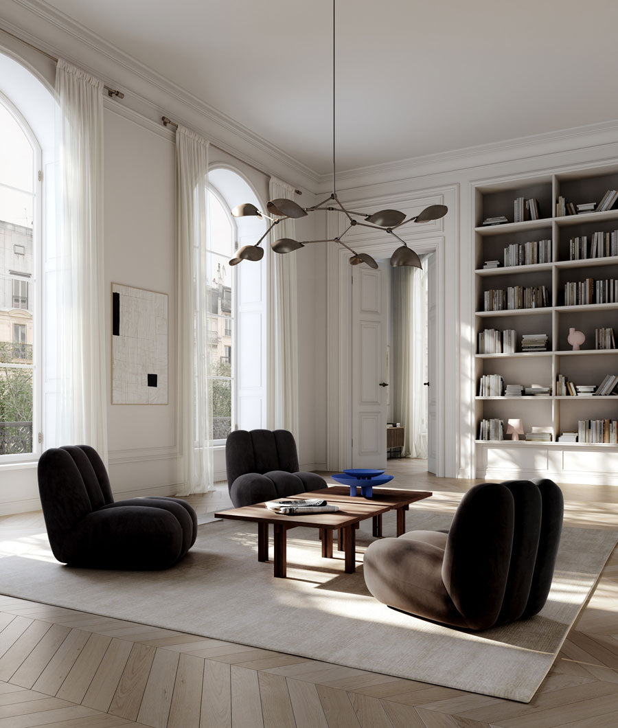 A modern living room with four dark upholstered Toe lounge chairs arranged around a wooden Plinth coffee table with Marrakech Blue Duck plates on a large cream rug. The room has high arched windows with white drapes, built-in bookshelves, light wood herringbone flooring, and a sculptural multi-arm Stingray Chandelier hanging from the ceiling.