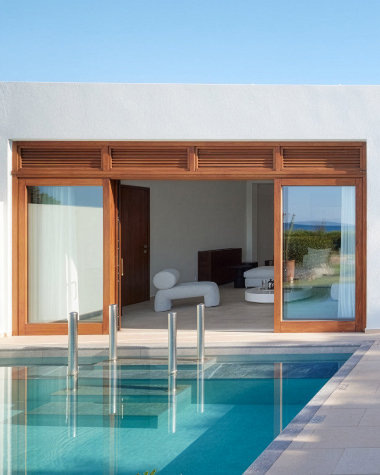 Modern architectural scene from the Amirands a Greotel resort. There is a swimming pool in the foreground, large wood-framed sliding glass doors opening to a minimalist interior showing a of white Foku Chair from 101 Copenhagen, and a clear blue sky above.