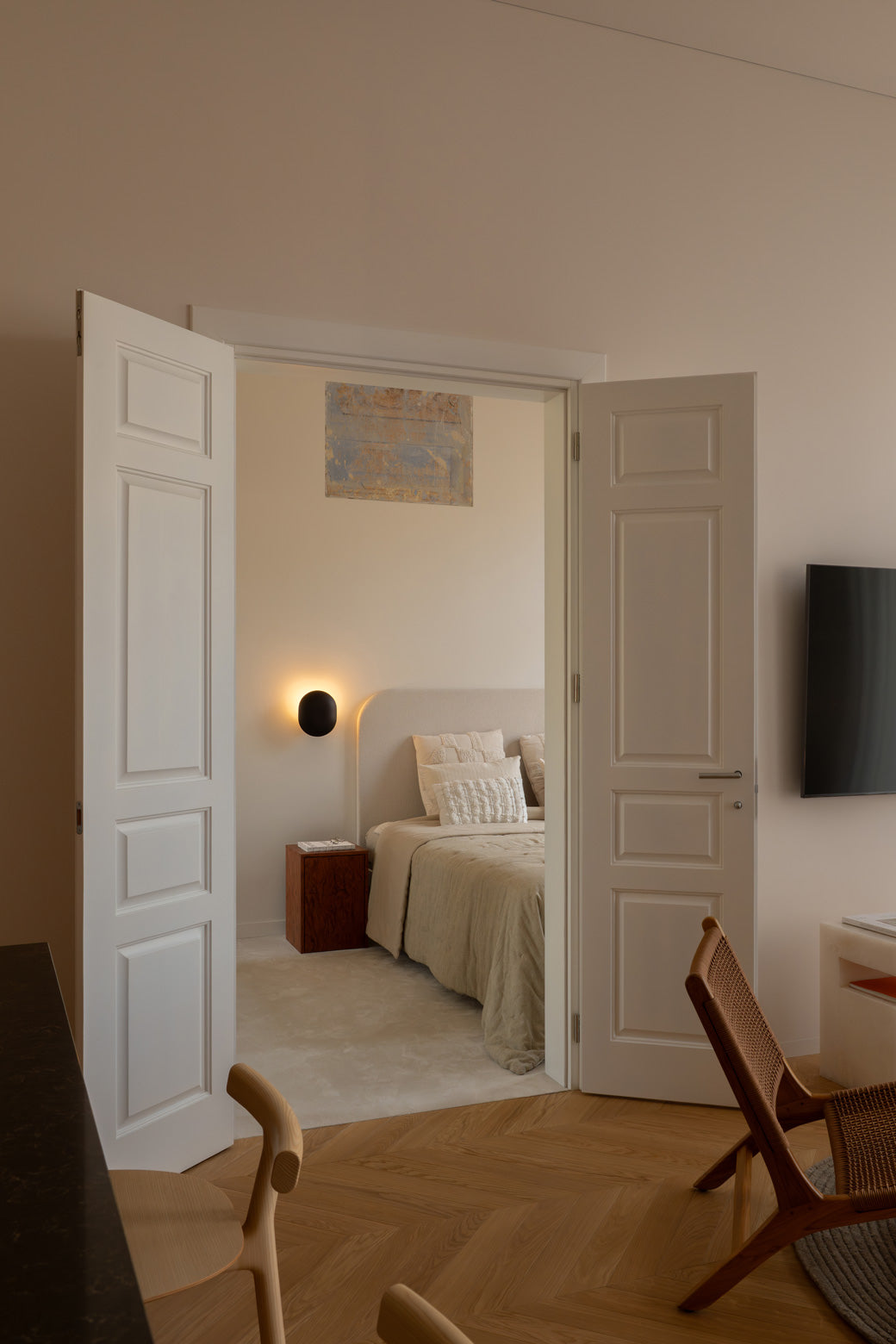 An open doorway in a modern bedroom setting, showing a neatly made bed with beige bedding and a lite Clam wall lamp. The room has warm beige walls, light wood flooring, and a wooden chair in the foreground.