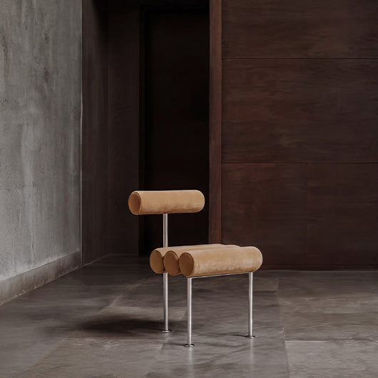 A modern minimalist Hiro dining chair with cylindrical beige velvet cushions on a chrome metal frame, set in a dark, industrial-style room with concrete floors and walls.