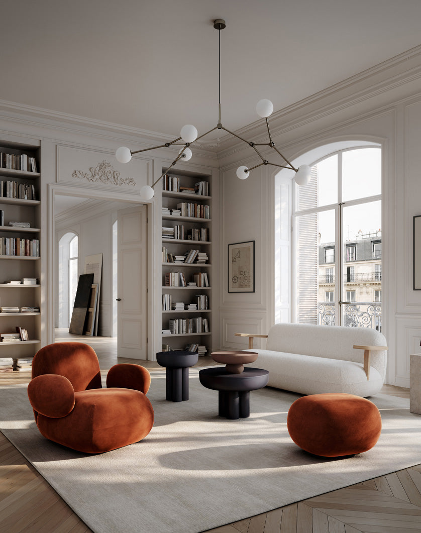 A luxury modern living room featuring a white Toe sofa, a rust-orange velvet armchair, with a matching ottoman, and dark round Crown coffee tables on a large cream rug. The room is styled with built-in bookshelves, arched windows, and the iconic 101 Copenhagen Drop chandelier Bulp.