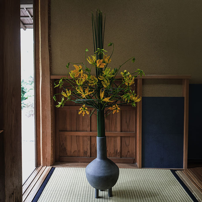 Decorative arrangement by Hikaru Seino in a grey Duck Vase on a tatami mat floor with wooden sliding doors in the background.