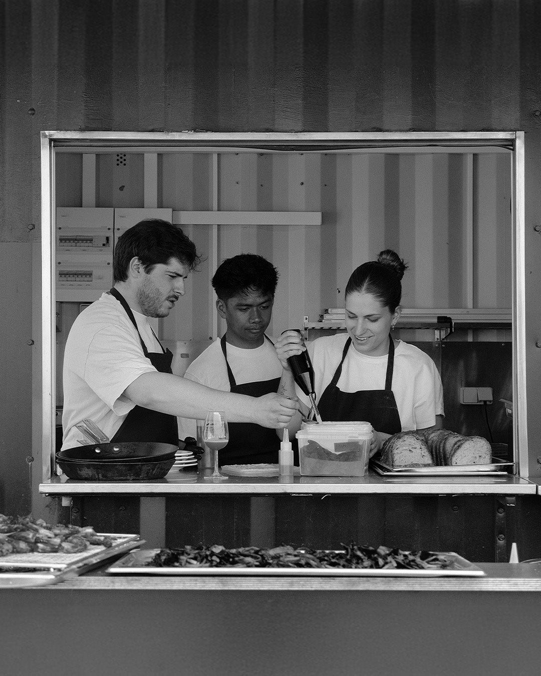 Black and white photograph of three chefs at a food service window, with trays of prepared food visible in the foreground.